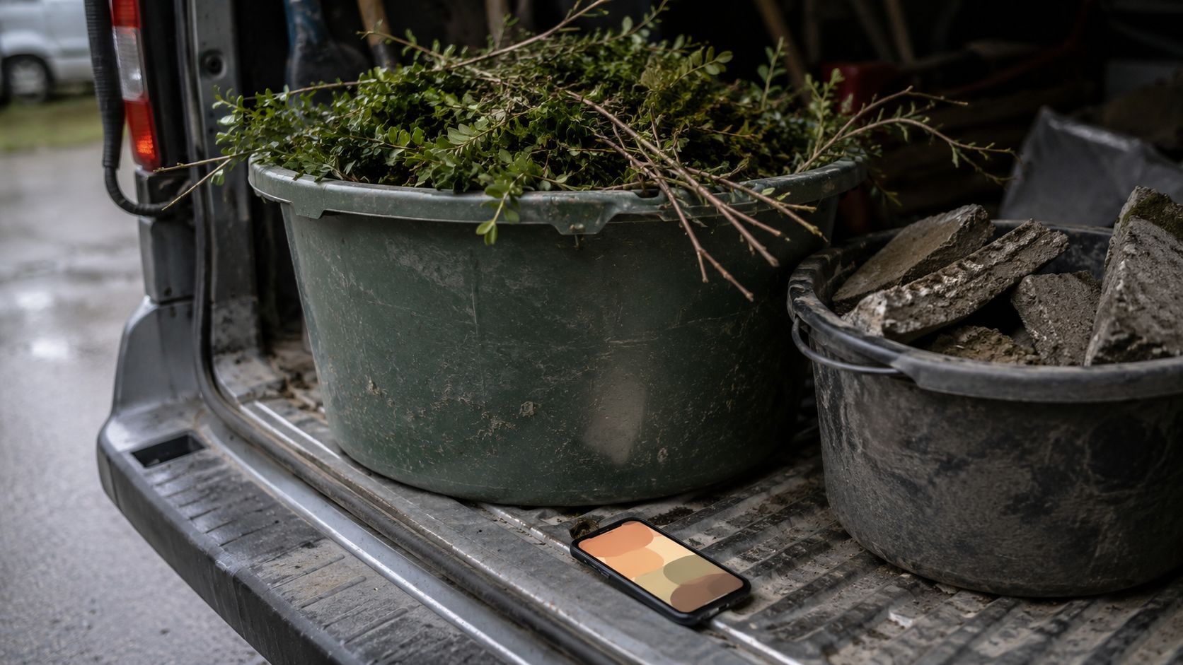 Groene kuip met snoeiafval en een grijze bak met puin achter in de bus, met een oplichtende telefoon op de rand